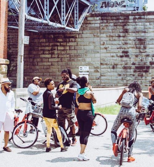 Projects 5 Friends on bikes gather in Cincinnati under a bridge on a sunny day.